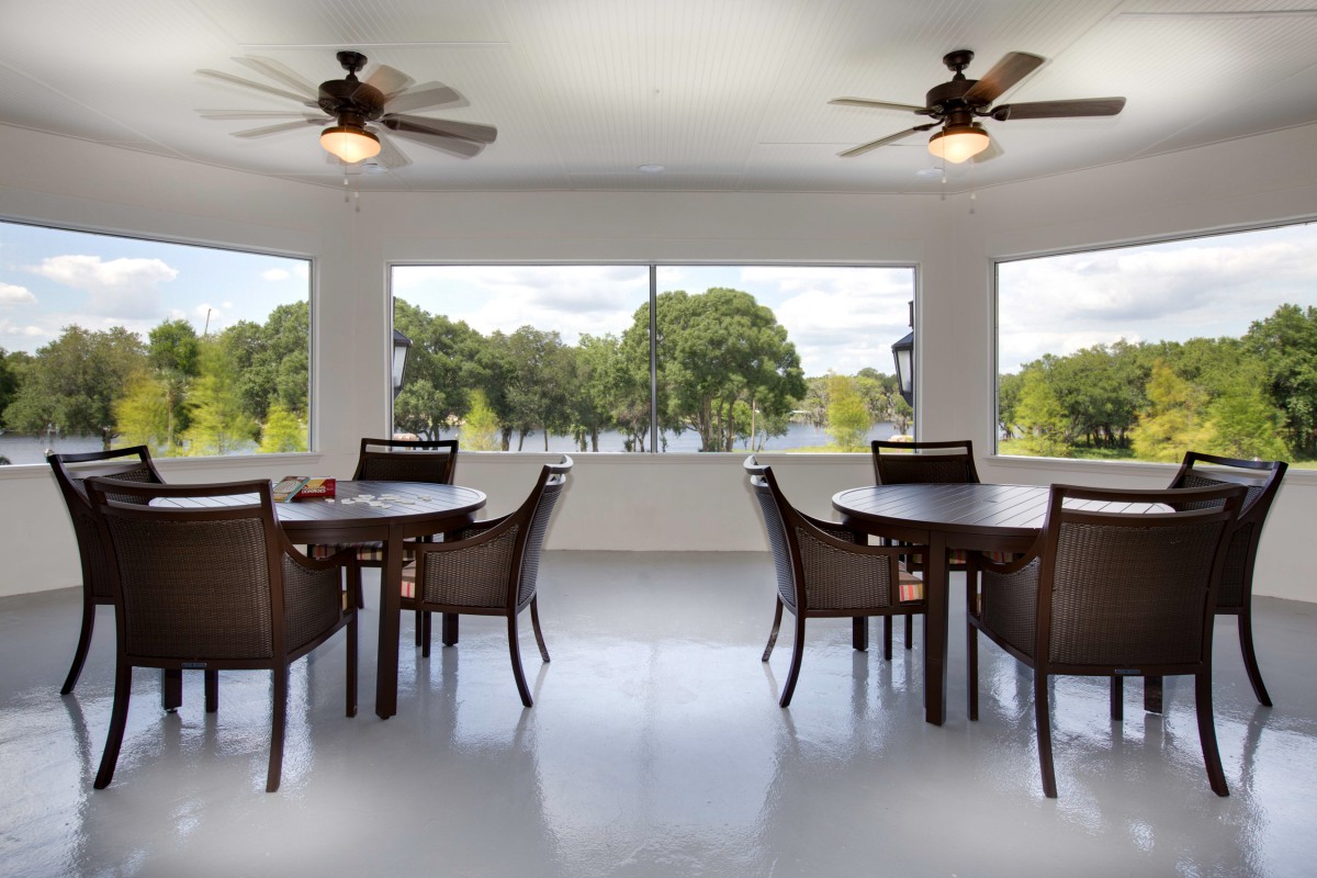 A bright dining area with multiple tables and chairs, featuring large windows and ceiling fans.
