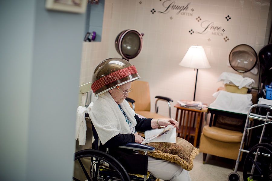 An elderly person sits in a wheelchair under a hair dryer, reading in a cozy salon.