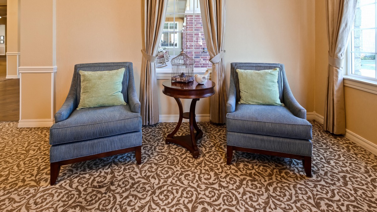 A cozy living room setup with two blue armchairs, a round wooden table, and a patterned carpet, framed by a window with elegant curtains.