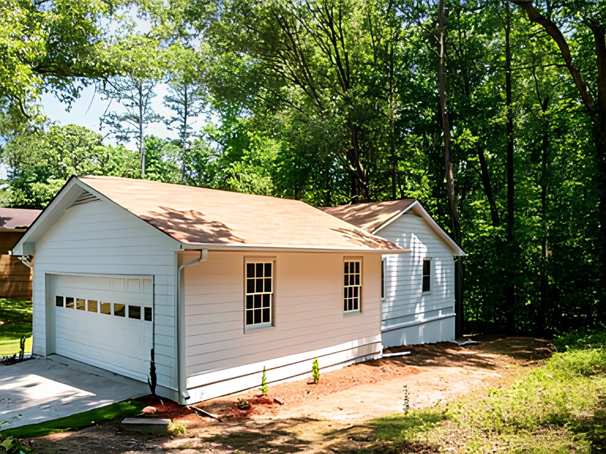 A small residential house with an attached garage surrounded by lush trees and basking in sunlight.