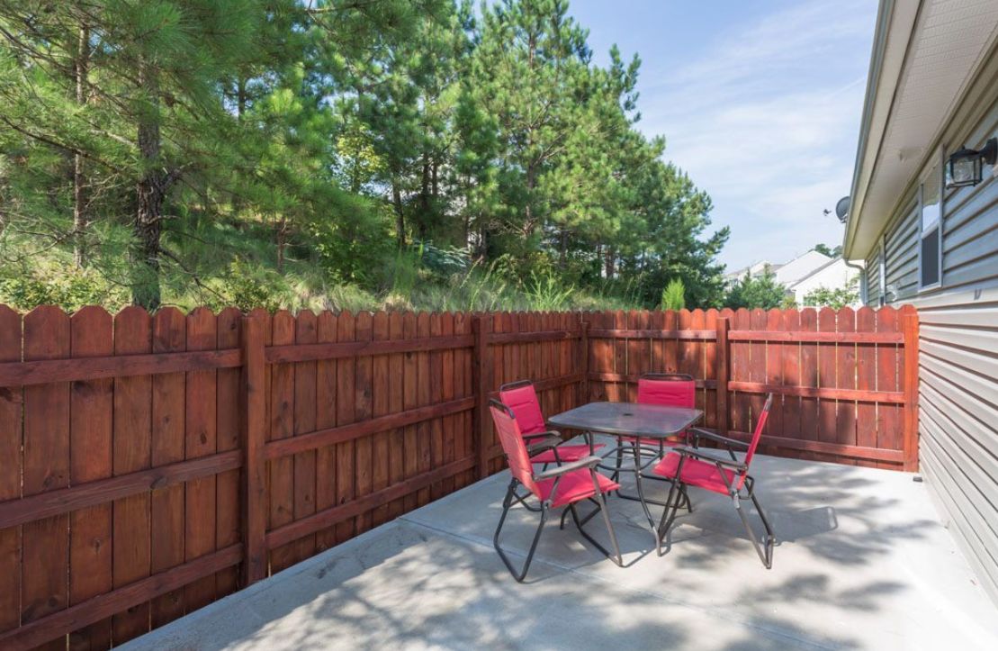 A cozy outdoor patio area with a table and red chairs, surrounded by a wooden fence and lush greenery.