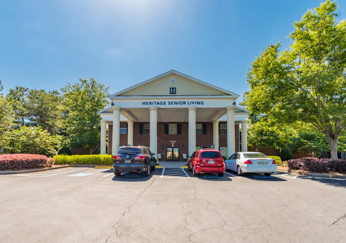 A front view of a building with large columns, surrounded by trees, and several cars parked in the lot on a sunny day.
