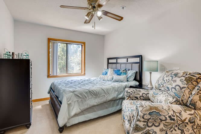 A cozy bedroom with a bed, armchair, ceiling fan, and large window.