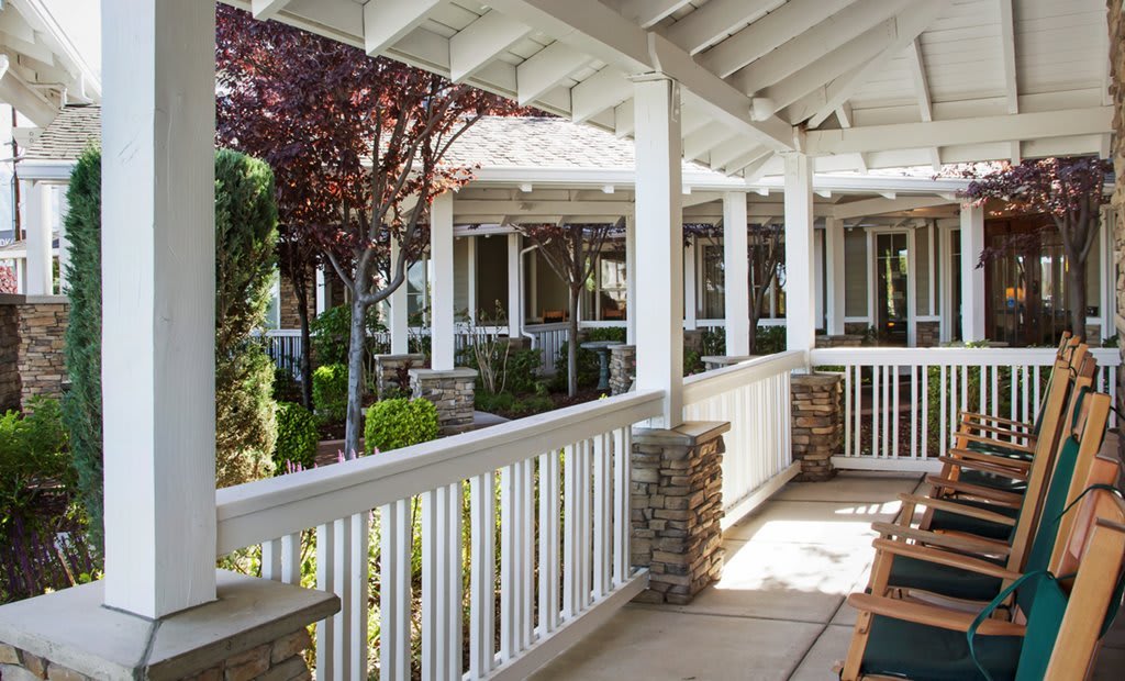 A cozy porch with white railings and columns, surrounded by a garden area with trees.