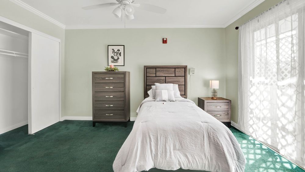 A neatly arranged bedroom with a single bed, dresser, and bedside table on a green carpet.