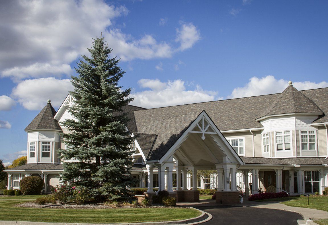 A large residential building with a steep roof and a tall tree in front, set against a backdrop of blue sky and clouds.