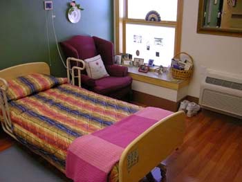 A cozy hospital room featuring a colorful bedspread, an armchair by the window, and various decorative items.