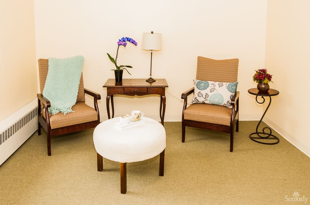 A cozy living room setup featuring two armchairs with cushions, a small side table with a lamp and orchid plant, and a round ottoman in the center.