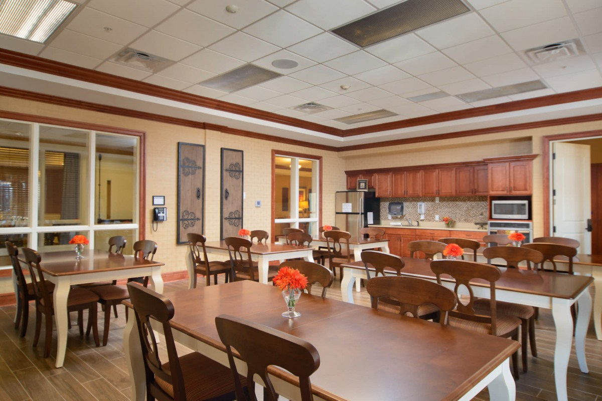 A communal dining area with wooden tables and chairs, featuring a kitchen area in the background and vases of flowers adorning the tables.