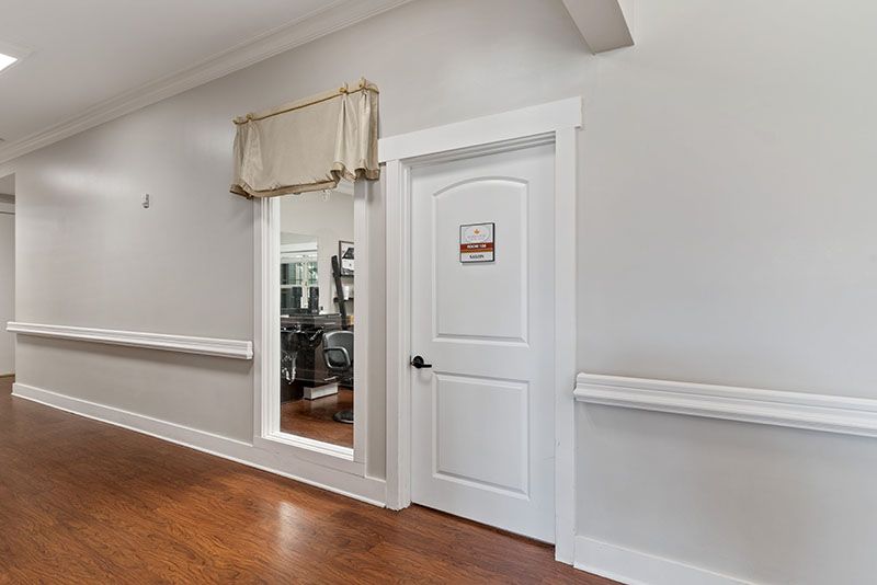 An interior hallway with a door leading to an exercise room, featuring a large mirror and hardwood flooring.