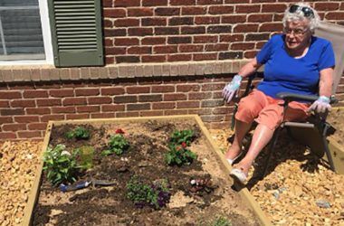 A woman sits next to a garden bed, watering the plants on a sunny day.