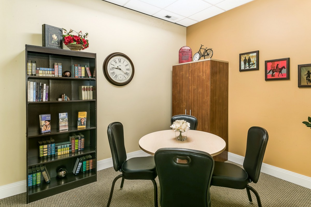 A small office meeting room with a round table, chairs, and a bookshelf.