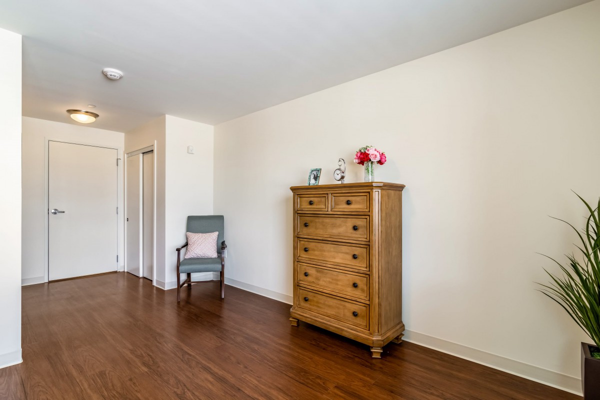 A minimalist room featuring a wooden chest of drawers and an upholstered chair on a hardwood floor.