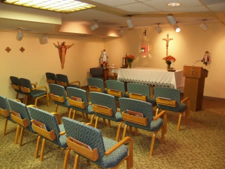 A small chapel with rows of chairs facing an altar, adorned with a cross and crucifix.