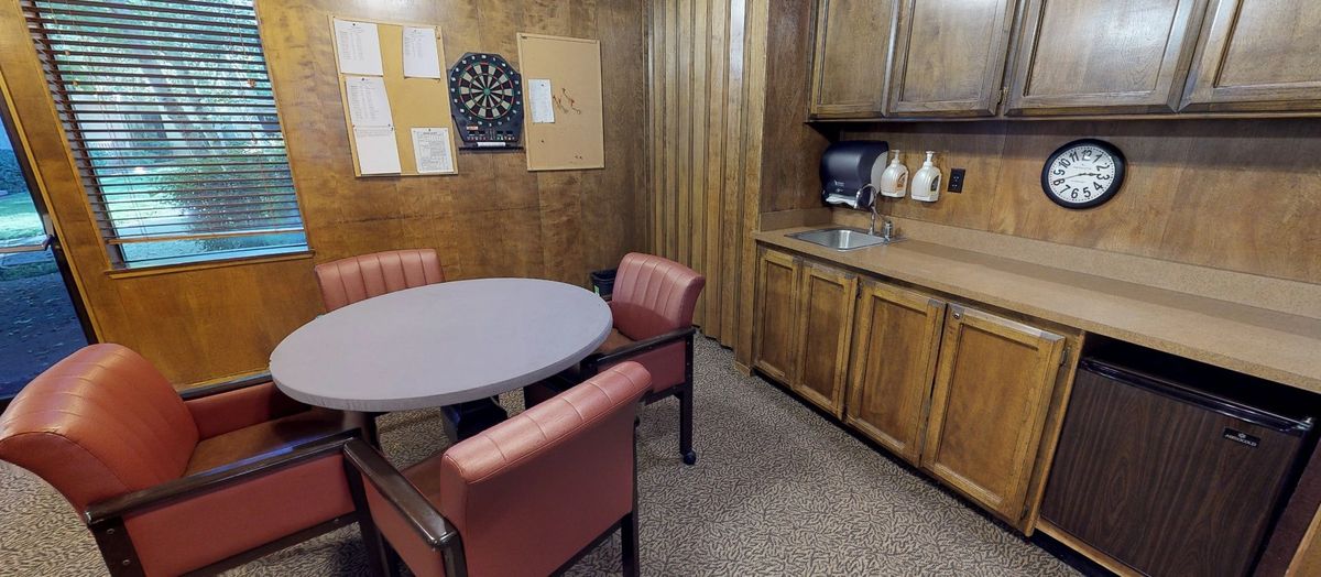 A small office break room with a round table, four chairs, wood paneling, and kitchen amenities.