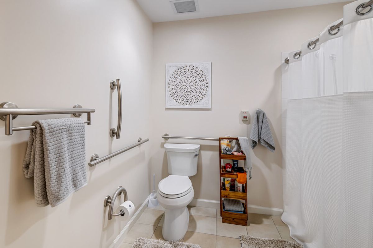 A neatly organized bathroom featuring a toilet, towel racks, a grab bar, and a small bookshelf with various items.