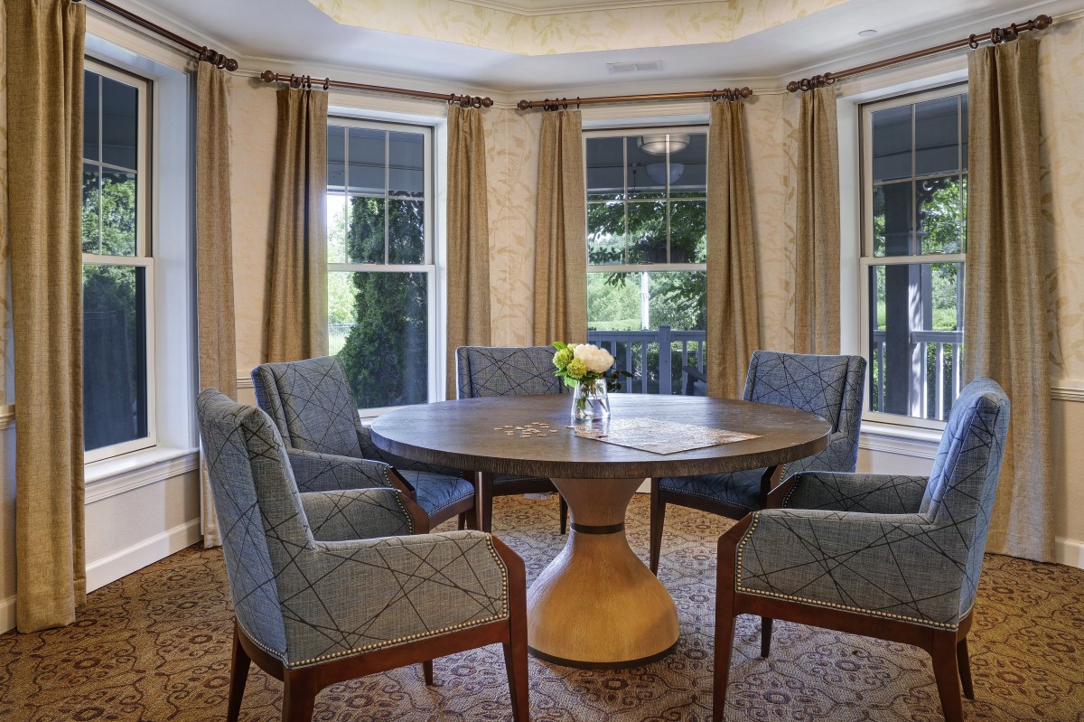 A cozy dining room featuring a round table surrounded by upholstered chairs and flanked by large windows with curtains.