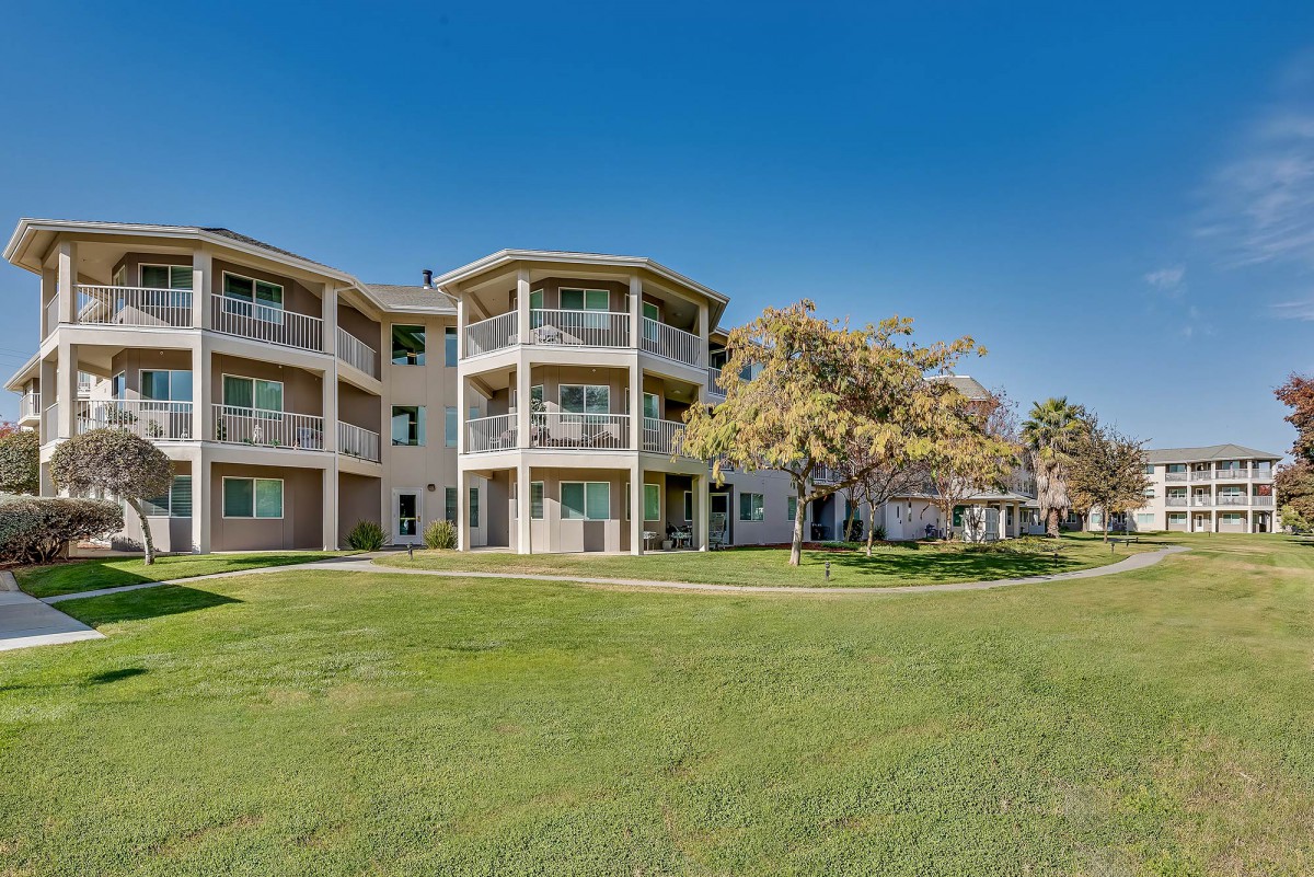 A residential apartment building with balconies, surrounded by a well-maintained lawn and trees under a clear blue sky.