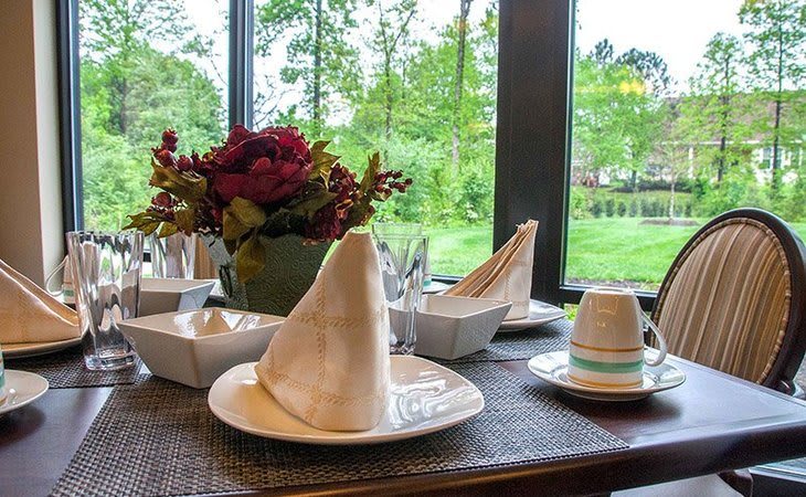 A beautifully set dining table with folded napkins, flowers, and a scenic window view.