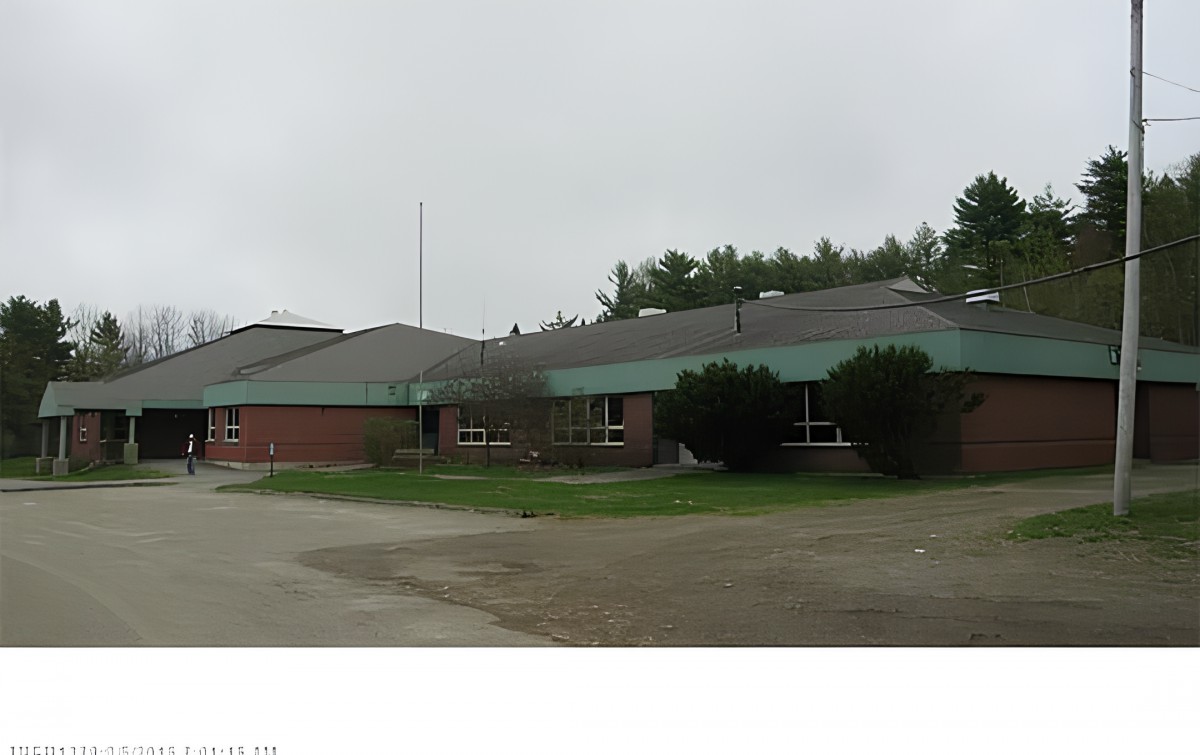 A single-story building with a sloped roof, possibly a school, surrounded by trees and overcast skies.