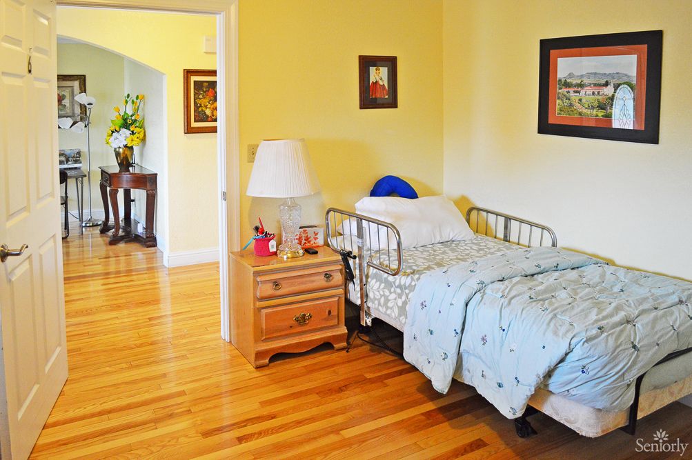 A cozy bedroom featuring a hospital bed with light blue bedding, a wooden nightstand with a lamp, and framed artwork on the walls.
