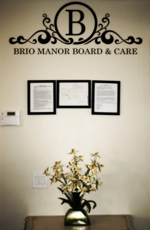 Framed certificates on the wall of Brio Manor Board & Care with a decorative plant below.