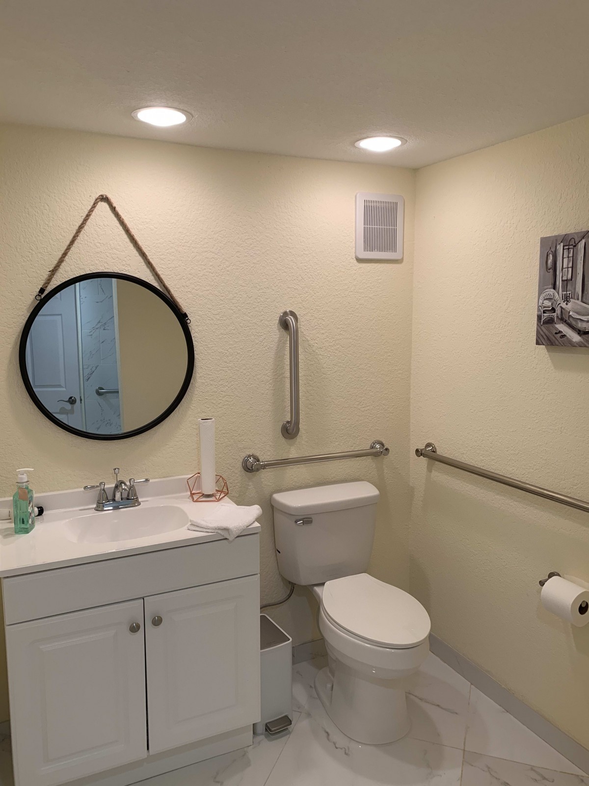 A modern bathroom with a round mirror, sink, and toilet featuring safety grab bars.