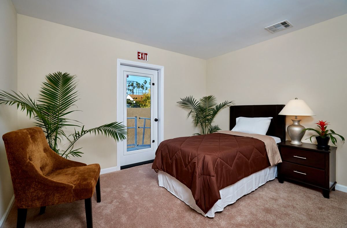 A cozy bedroom with a single bed, tropical plants, and a door marked with an exit sign.