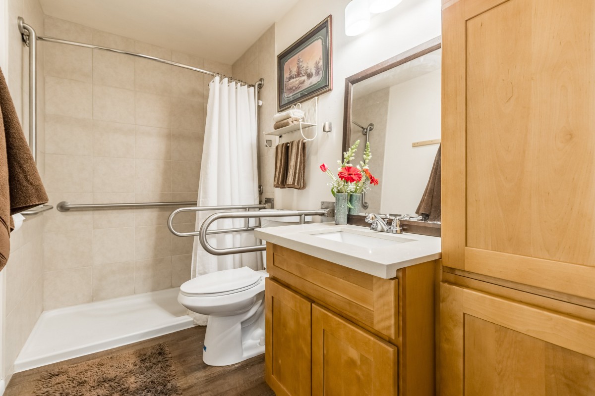 A well-organized bathroom featuring a sink with wood cabinets, a mirror, and a shower with a grab bar.