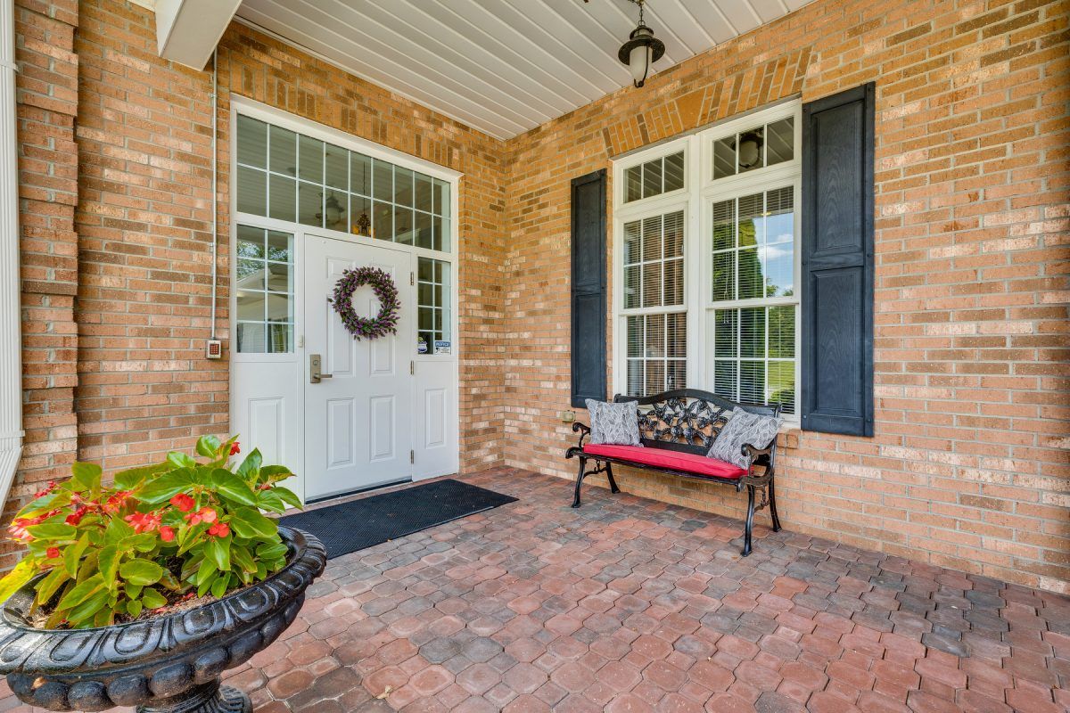 A welcoming front porch with a white door adorned with a wreath, brick walls, and a cozy bench beside potted plants.