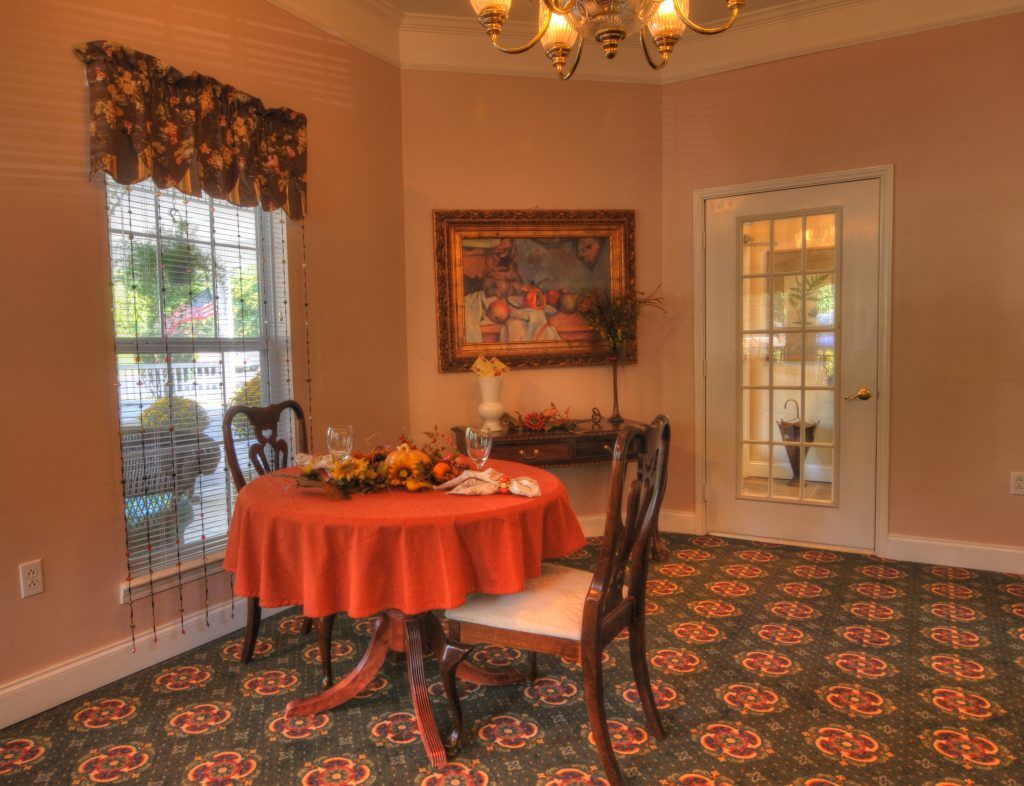 A cozy dining room with a round table covered by an orange tablecloth, surrounded by elegant chairs and adorned with a decorative carpet.