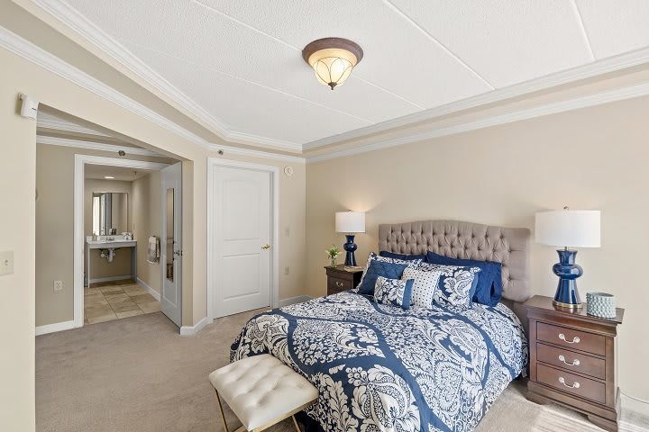 A cozy bedroom with elegant blue and white bedding, featuring a tufted headboard, nightstand with a lamp, and an en-suite bathroom.
