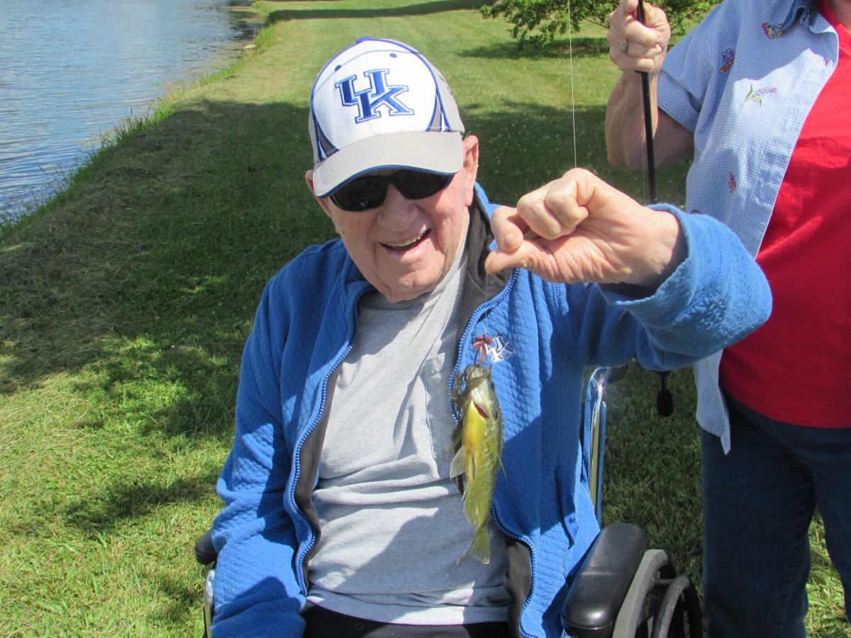 An elderly man in a wheelchair, wearing a hat and sunglasses, proudly holds up a small fish he caught by the water.