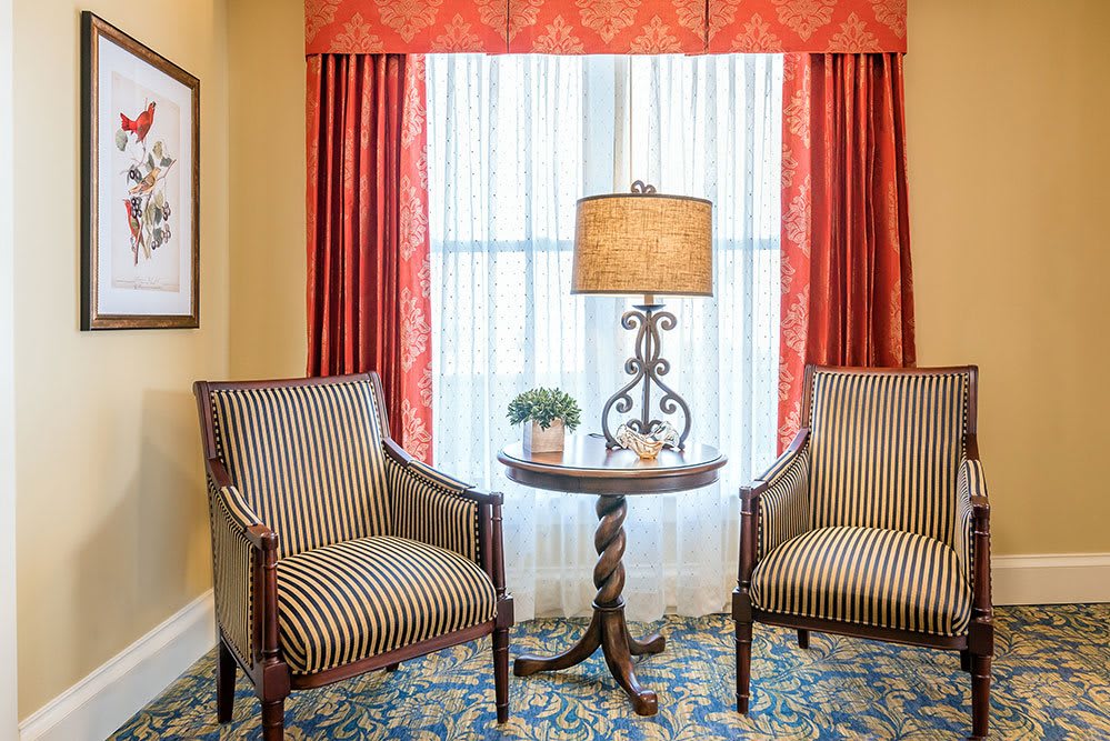 A cozy living room corner with two striped chairs, a round table, and a decorative lamp in front of a window with red curtains.