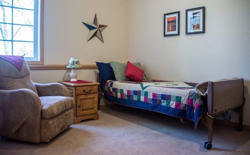 A cozy bedroom features a single bed with a colorful comforter, a few pillows, and a side table with a lamp beside an armchair. The wall is decorated with star-themed art and framed pictures.