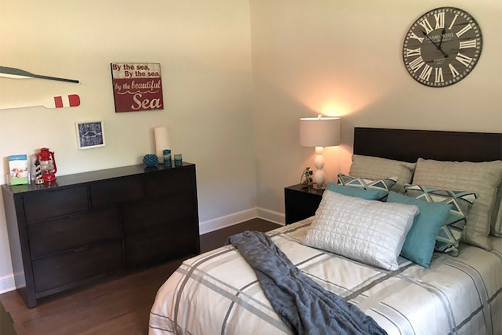 A cozy bedroom with a neatly made bed, decorative pillows, and a lamp on the nightstand. Wall art and a clock adorn the walls, creating a welcoming atmosphere.