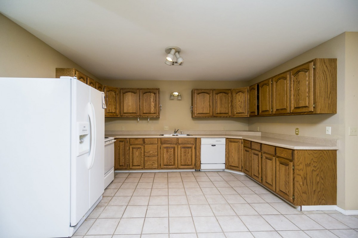 A spacious kitchen with wooden cabinets, a white refrigerator, and a dishwasher, featuring a tiled floor.