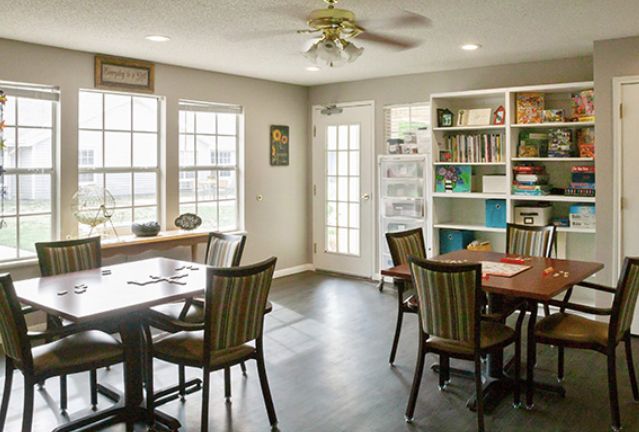 A cozy dining area with several tables and chairs, complemented by a bookshelf and large windows that allow natural light to brighten the space.
