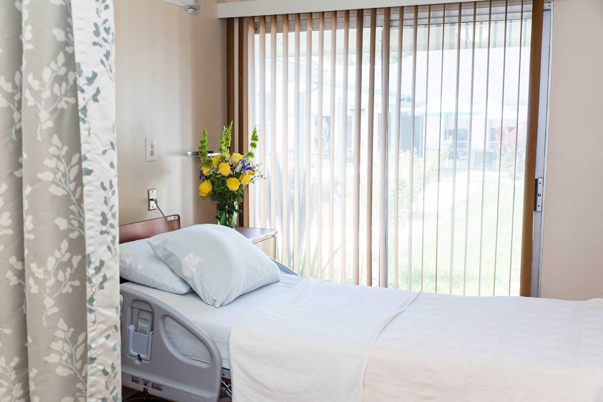 A bright hospital room with a neatly made bed and flowers by the window.