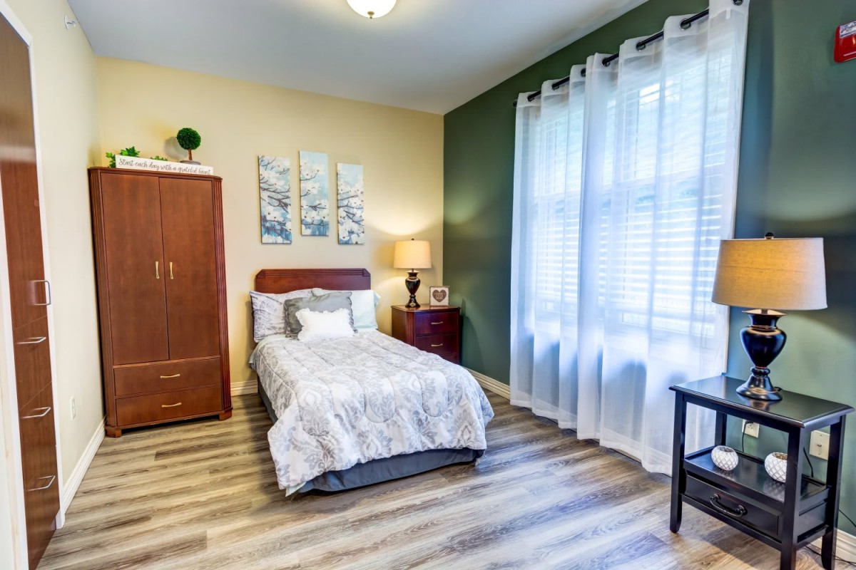A cozy single bedroom with modern decor, featuring a large window with sheer curtains and wooden flooring.