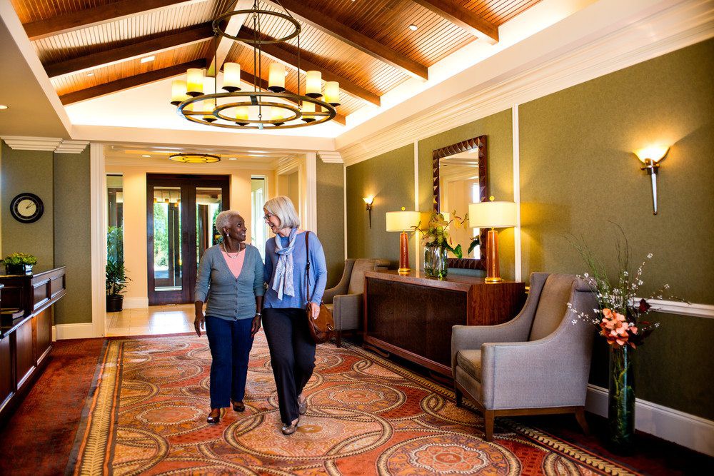 Two people walk through a well-decorated hallway with comfortable seating and warm lighting.