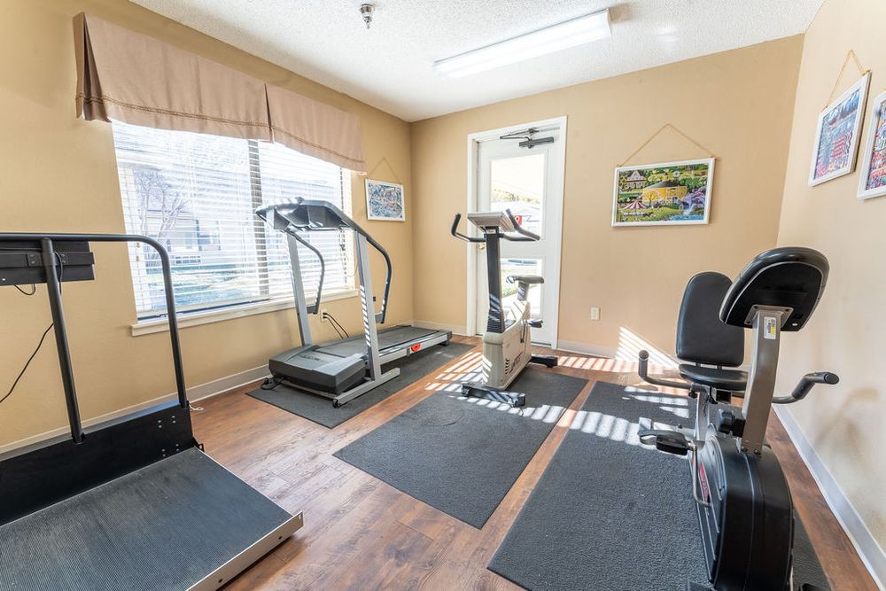 A small gym room equipped with treadmills and other exercise machines, well-lit by natural light from large windows.