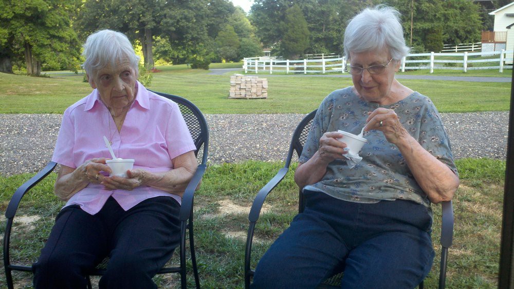 Two elderly women enjoying ice cream while sitting outside in a garden setting.