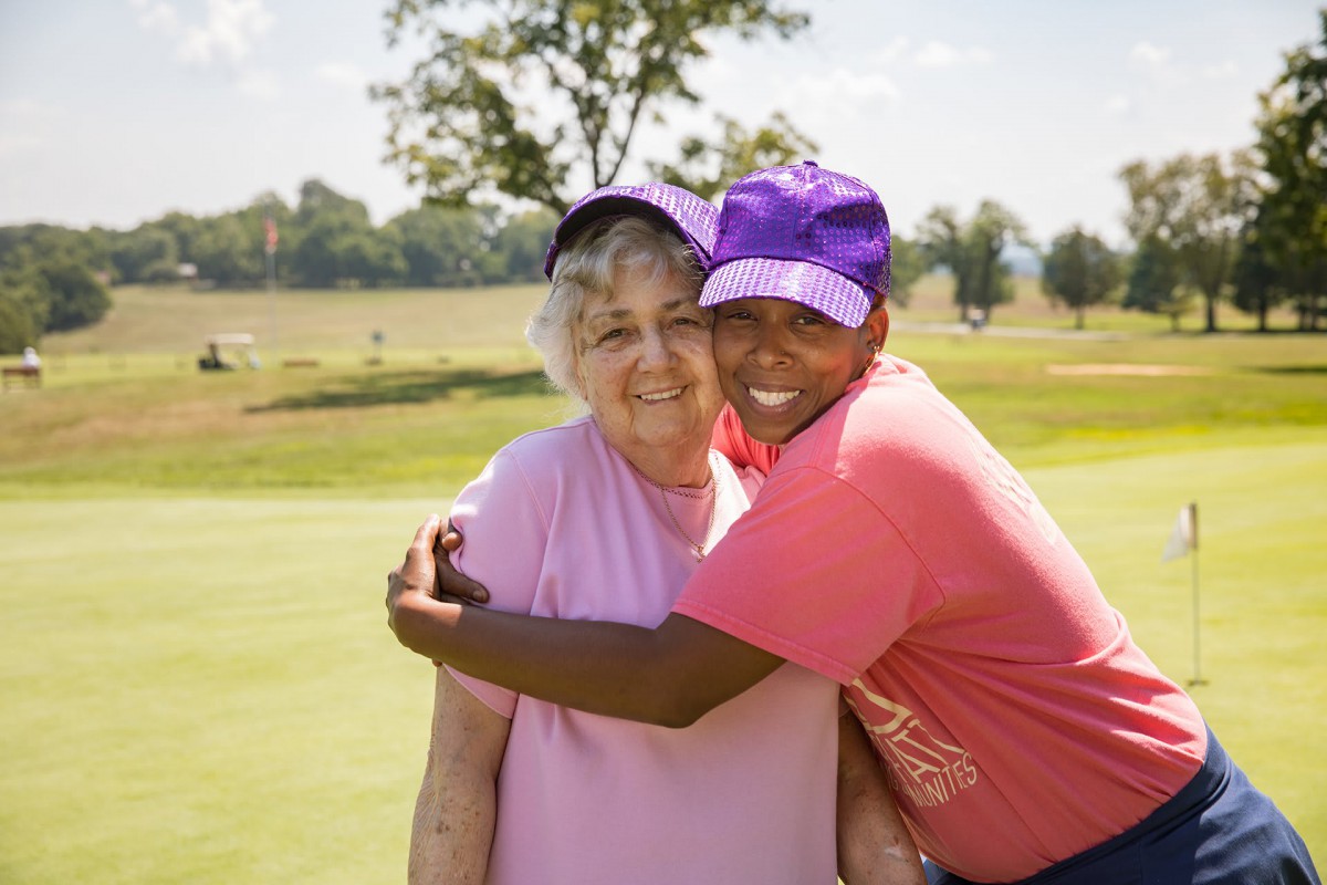 Two people hugging and smiling outdoors on a sunny day, wearing hats.