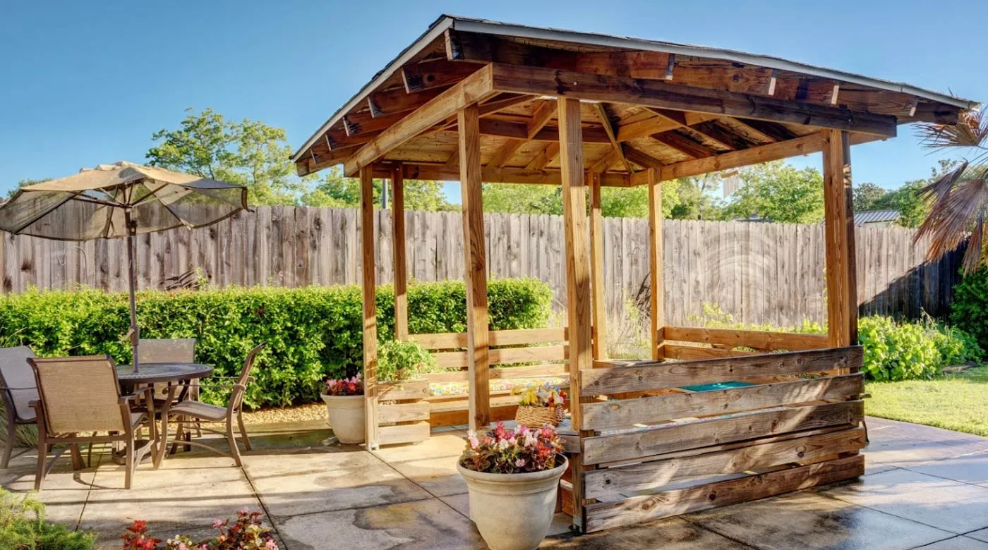 A sunny backyard featuring a wooden gazebo surrounded by potted plants.