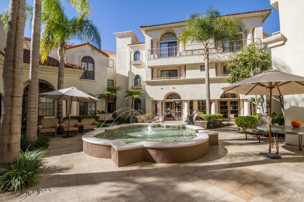 A sunny courtyard with a fountain and outdoor seating surrounded by Mediterranean-style apartment buildings.