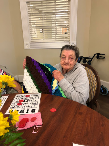 An elderly woman proudly displays her colorful crochet project while enjoying a game of bingo with friends.