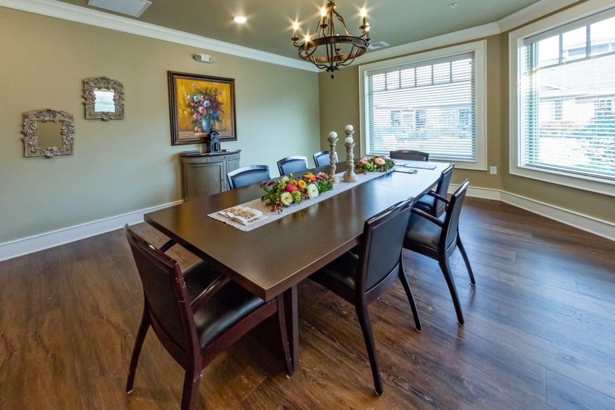 A well-decorated dining room featuring a large wooden table surrounded by chairs, with a chandelier overhead and a bright window lighting the space.