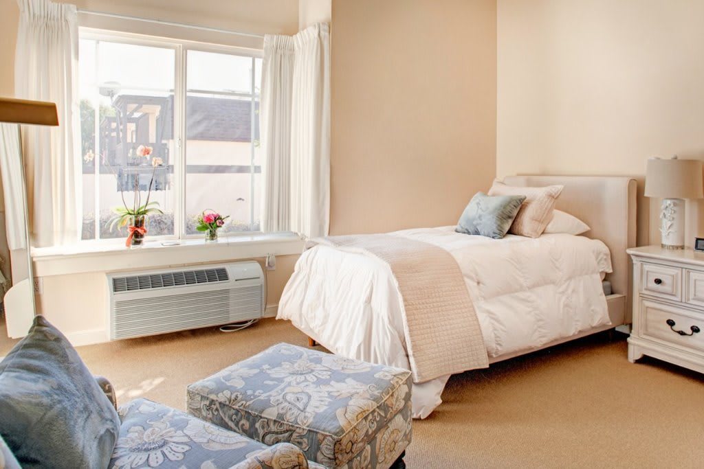 A cozy bedroom featuring a single bed with light-colored bedding, soft cushions, and a large window allowing natural light to fill the space.