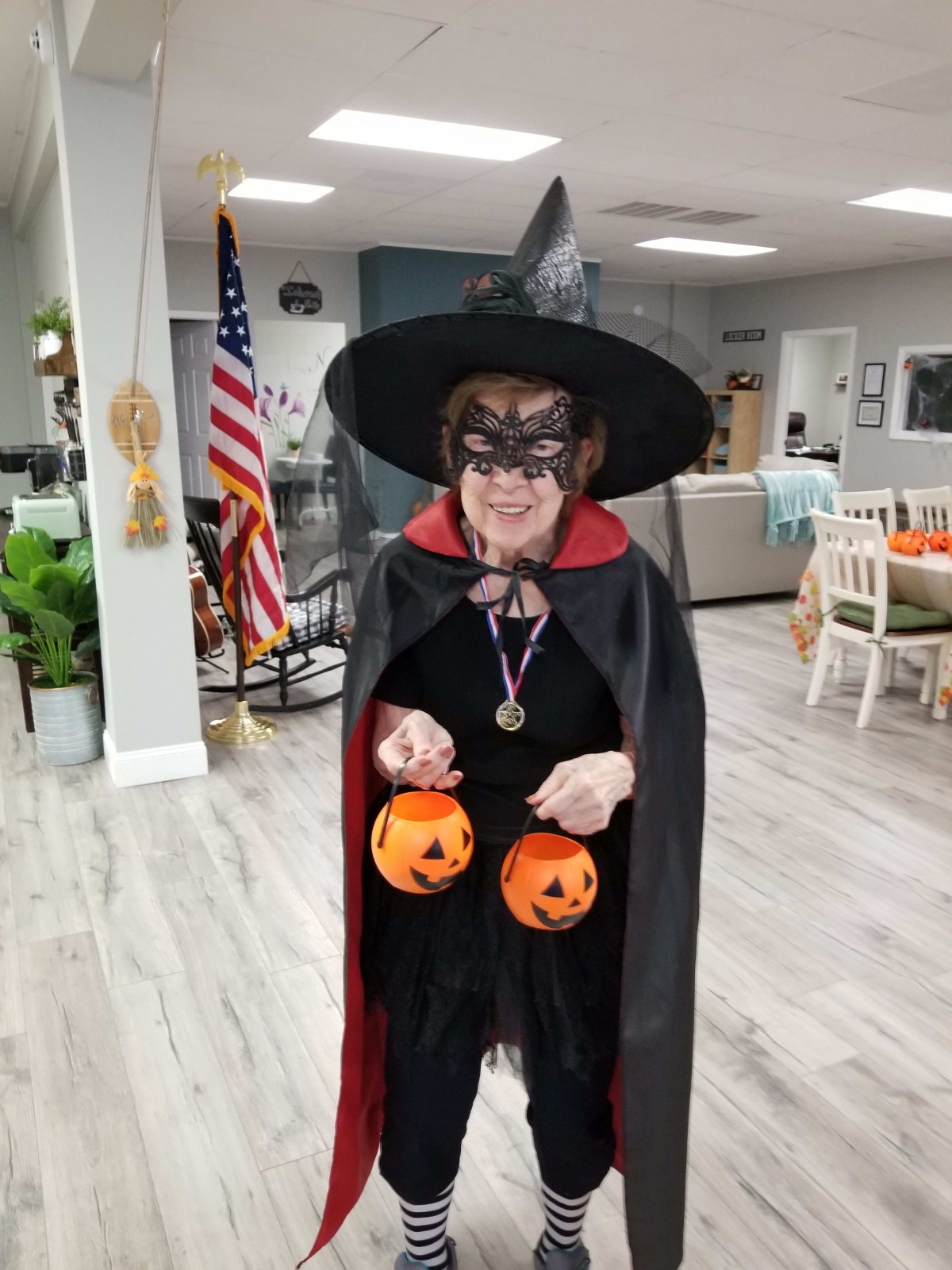 A person dressed as a witch for Halloween, holding two pumpkin-themed buckets indoors.